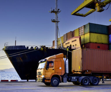 Truck carrying forty-foot container leaving port terminal with ship and quay crane on the background. Seaport operation activities, container shipping, and logistics.