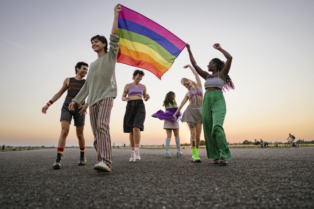 View of a group of young people holding a rainbow flag together, half-dancing and smiling along a path