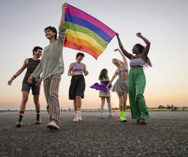 View of a group of young people holding a rainbow flag together, half-dancing and smiling along a path